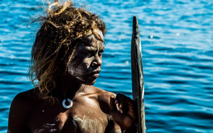 indigenous pacific islands boy holding a shield with deep blue water rippling behind him