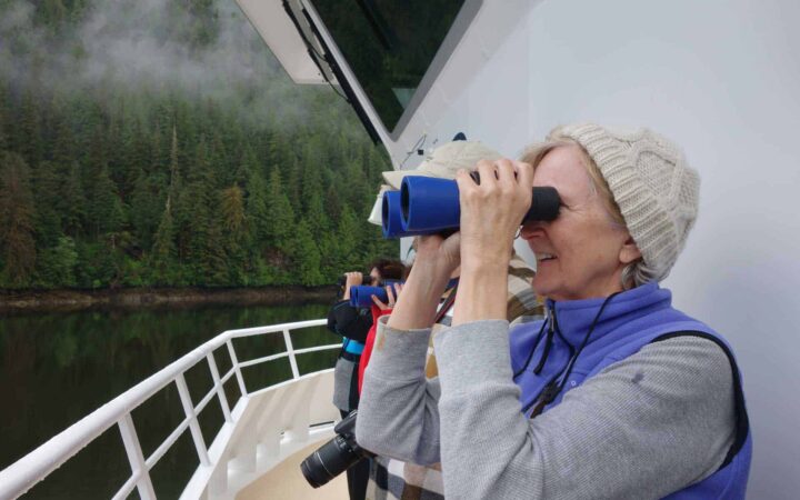 woman on the front of an alaska small ship holding binoculars looking out over the bow on a misty day with green forest in the background
