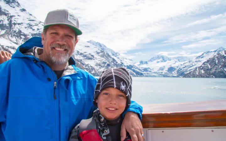 adventuresmith explorations president and founder todd smith and his son smiling on the bow of an alaska small ship with large snowy mountains behind them