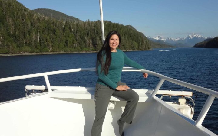 adventuresmith explorations adventure specialist leslie camacho sits on the bow of a luxury small ship in alaska on a sunny day with mountains in the distance