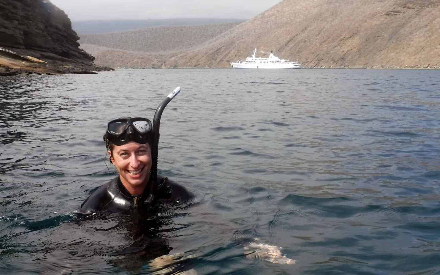 smiling woman with a mask and snorkel pops her head out of water while the legend galapagos small ship anchors in the distance behind her