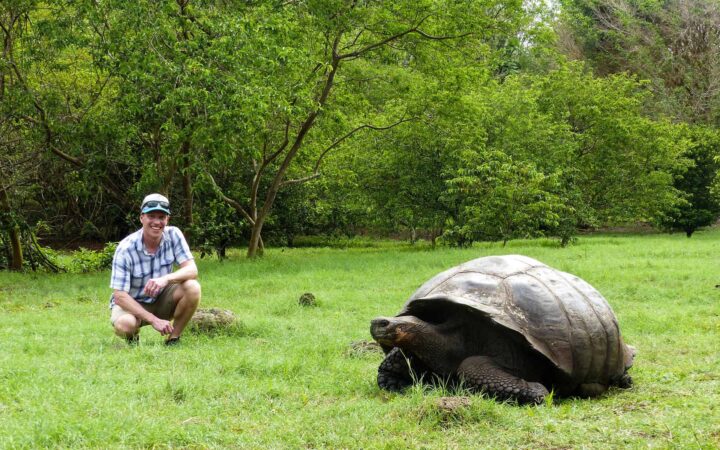 man sitting smiling next to a tortoise on a galapagos land tour with lush green grass and foliage around them