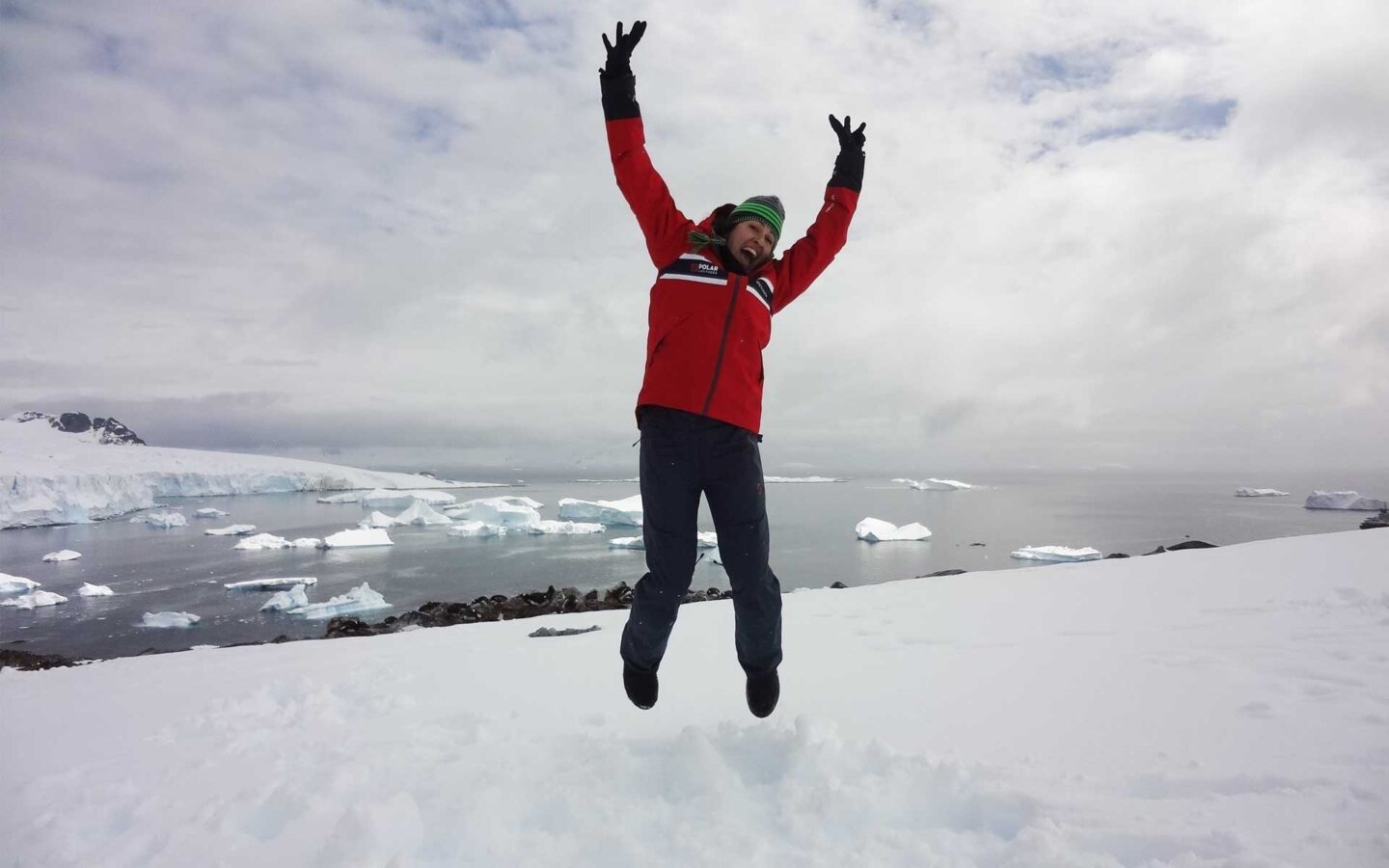 female solo traveler in a red jacket jumping with her arms up on an antarctica mountain with the water behind her