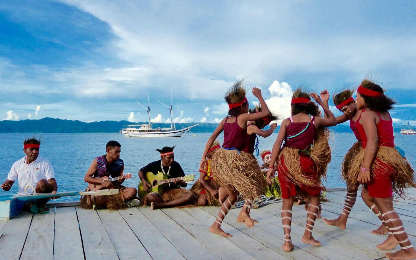 indigenous indonesian women wearing grass skirts dance on a dock with a luxury small ship anchored in the distance