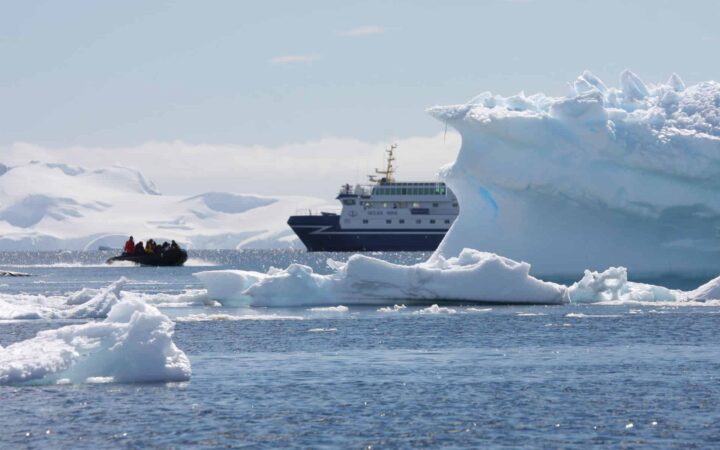 the front of an antarctica expedition ship peaks out from behind an iceberg while a zodiac raft carries adventure travelers away from it on a sunny day