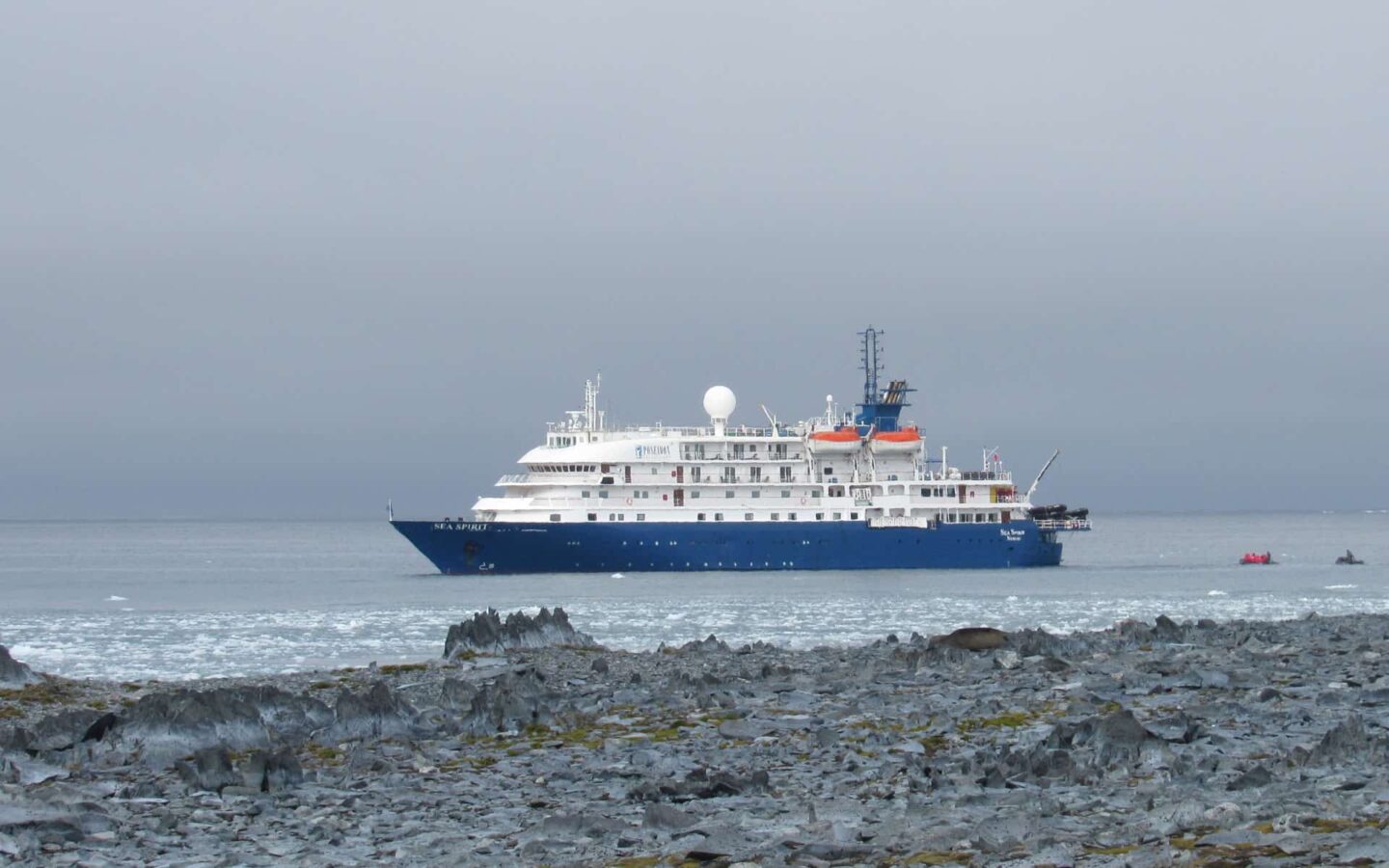 antarctica expedition ship cruising on calm water with travelers in zodiac boats behind it and a rocky shoreline in the foreground