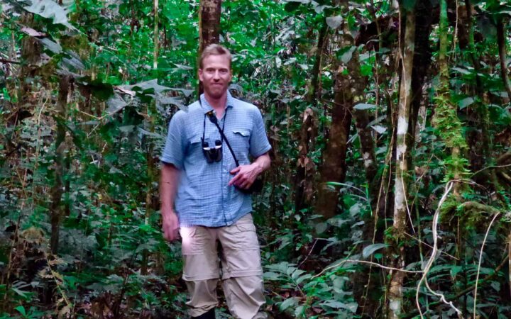 adventure traveler with a blue shirt and binoculars around his neck standing smiling in the amazon rainforest