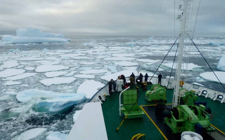 adventure travelers standing on the bow of an antarctica expedition ship with sea ice patches in front of them and snow mountains in the distance