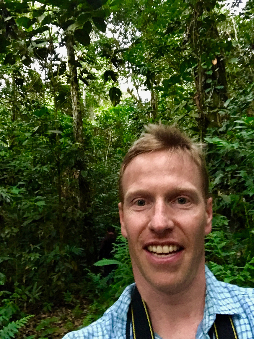 Man in blue and white collared shirt smiling with a jungle in the background.