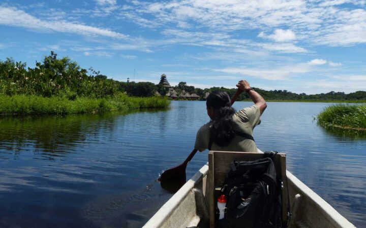 Amazon guide padding a canoe to an Amazon wilderness lodge on a river with green foliage on each side.