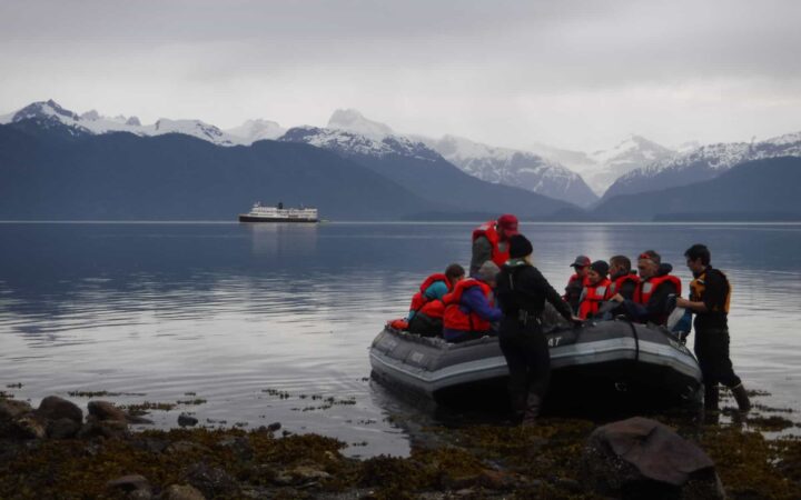 group of adventure travelers wearing red jackets load a zodiac skiff on shore with an alaska small ship anchored in the distance on an overcast day