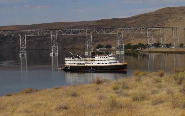 pacific northwest small ship anchored in the columbia river with a bridge behind it and brown grassy fields around it