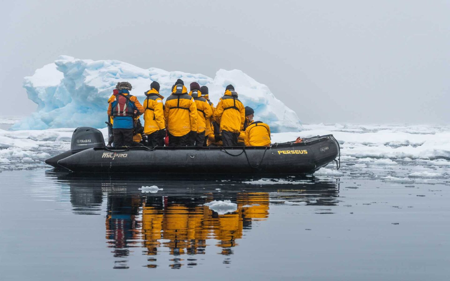 antarctica travelers in yellow jackets cruise in a zodiac skiff with an iceberg behind them on an overcast day