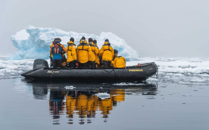 antarctica travelers in yellow jackets cruise in a zodiac skiff with an iceberg behind them on an overcast day