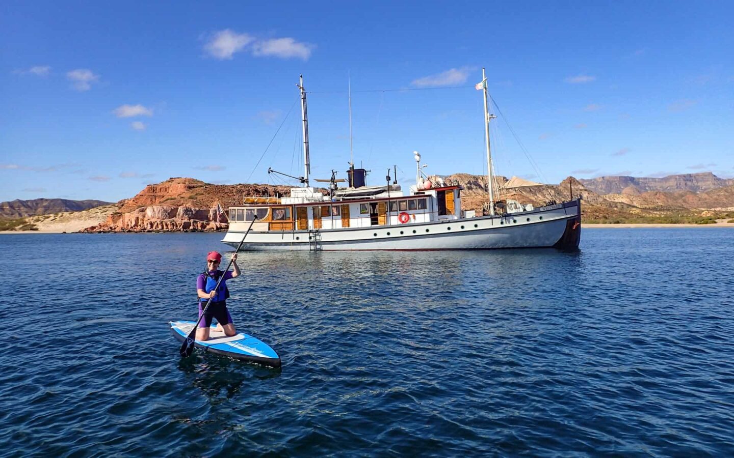 A Baja traveler paddleboarder in front of a small ship with desert island in the background.