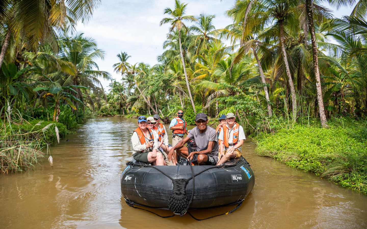 Black Zodiac boat with Panama small ship cruise travelers floats on muddy river under swaying palm trees.