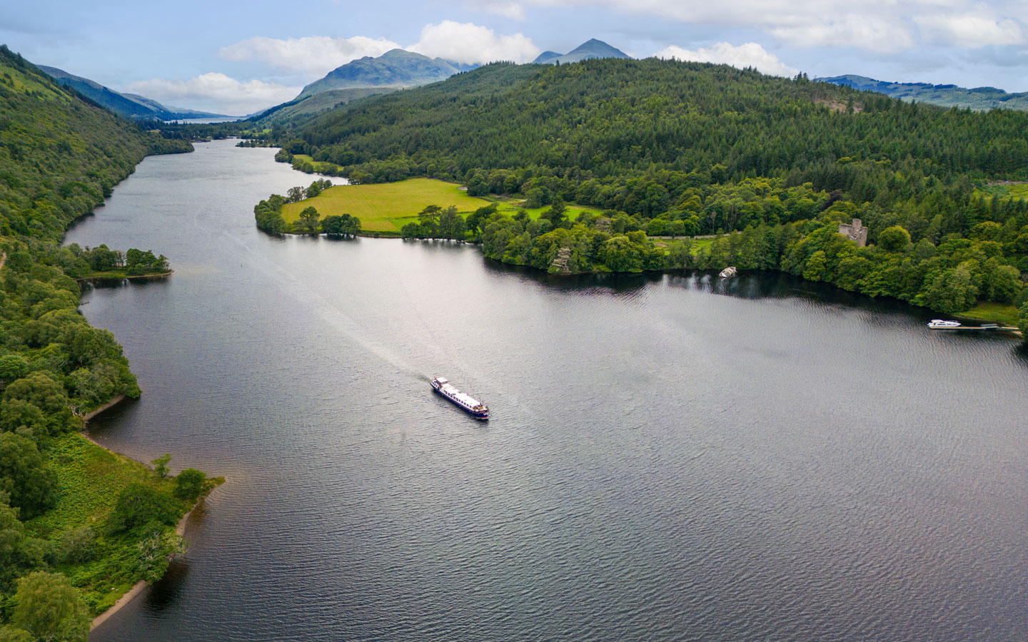 Small hotel barge motors through serene landscape of green hills & partly cloudy skies on small ship cruises in Northern Europe.