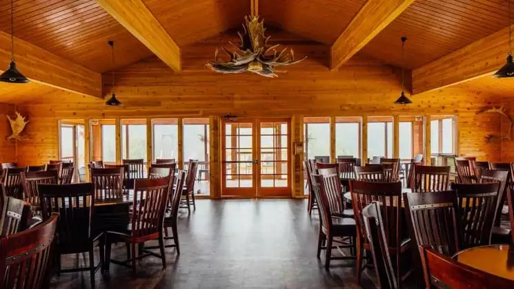 Large dining room with round wooden tables, wooden chairs, wall of windows, and large hanging light with moose antlers at Denali Backcountry Lodge in Kantishna, Alaska