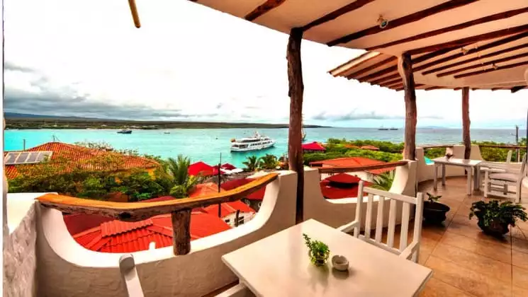 a table and chairs sitting on the balcony of the Angermeyer Waterfront Inn galapagos hotel with the ocean and a small ship in the background