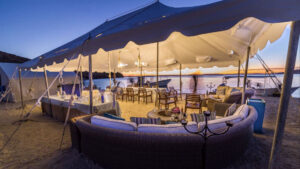 Open-air dining area under a white tent on a beach in Baja, with dining room table, candelabra & lounge area at Camp Cecil de la Isla