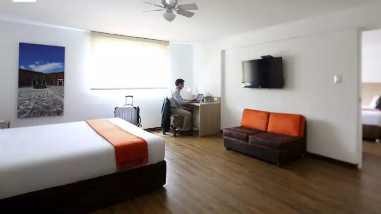 A person works on wifi on hiis computer at the desk inside a traditional room at the Casa Andina Standard Arequipa, a comfortable Peruvian hotel. The room has a full bed, television, and couch.