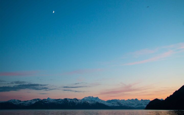 overlooking a channel in alaska at night with snowy mountains in the distance and the moon high above