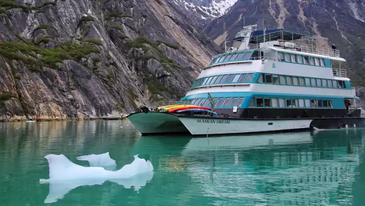 Alaskan Dream catamaran in Alaska with 3 blue & white decks, view windows throughout & sun deck, sitting beside iceberg bit.