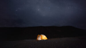 Lone yellow & white dome tent glows from within on a sandy beach under a starry sky at Camp Cecil de la Isla, Baja California.