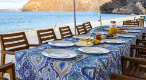 A long table is set with a blue print tablecloth, plates, and chairs for beachside dining at Camp Cecil de la Isla, where Espiritu Xantus Café is where fresh, healthy meals are prepared by the on-site chef.