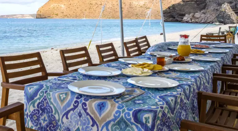 A long table is set with a blue print tablecloth, plates, and chairs for beachside dining at Camp Cecil de la Isla, where Espiritu Xantus Café is where fresh, healthy meals are prepared by the on-site chef.