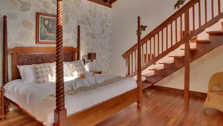 Four-post bed with wood detailing and local artwork adoring the walls, with stairs leading to a sunny balcony at Caves Branch Jungle Lodge in Belize