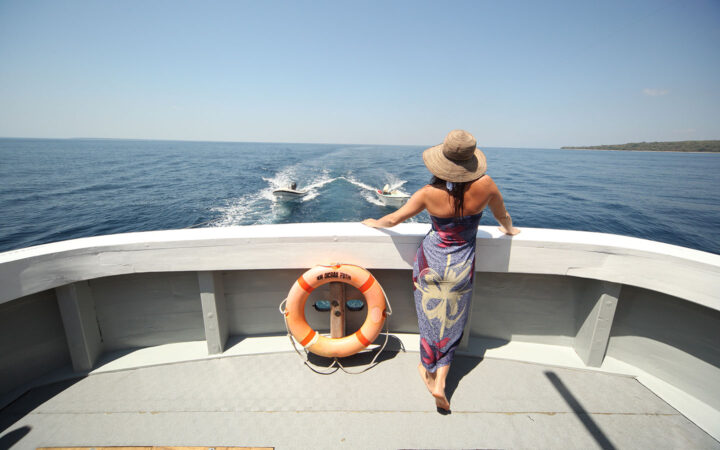A woman in a sarong and straw hat faces out to see on the stern of a small charter cruise yacht