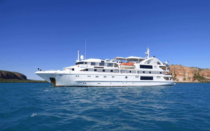 coral expeditions luxury small ship cruising on blue water on a sunny day with sheer cliffs behind it