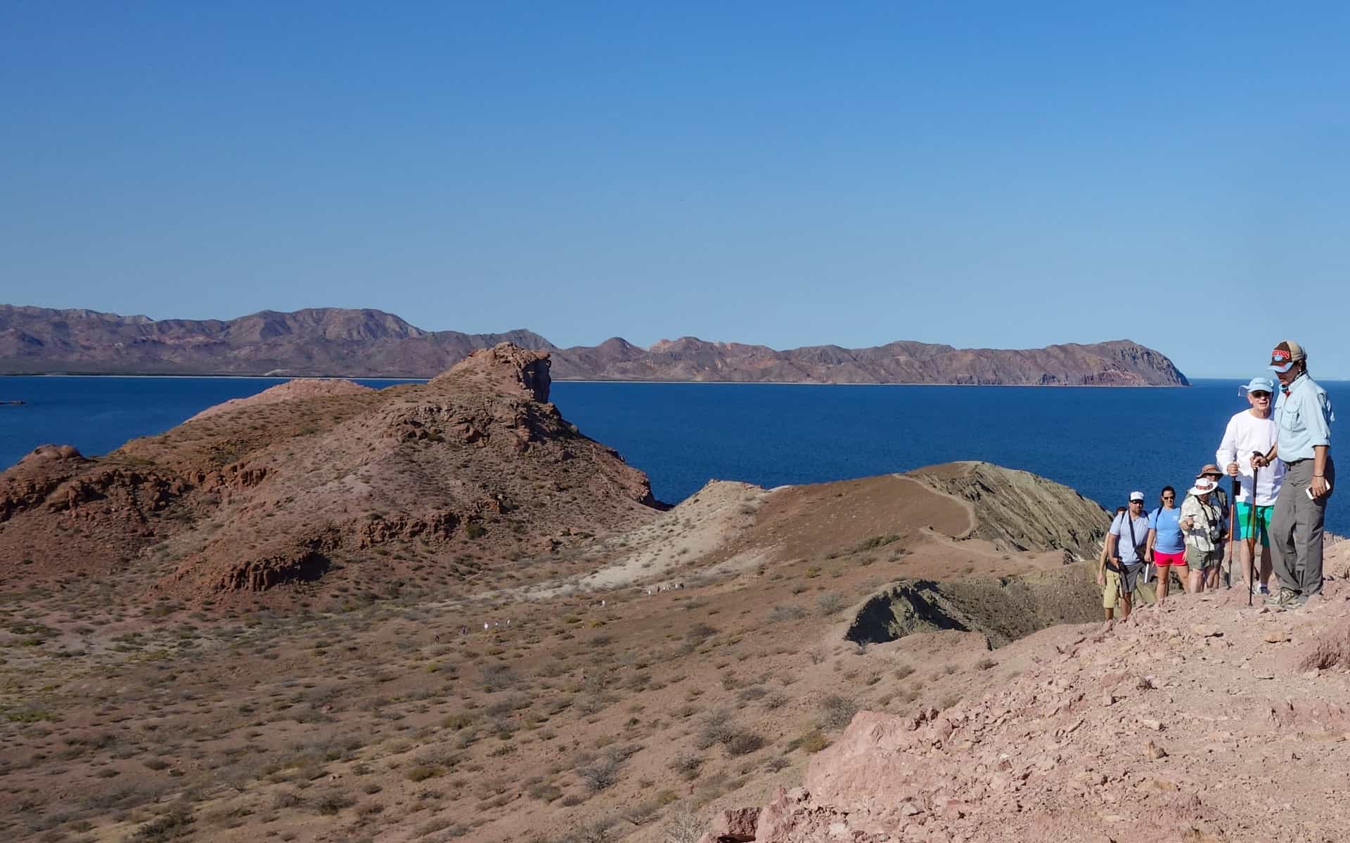 A group of travelers hiking up a desert lookout over the blue ocean with mountains in the background.