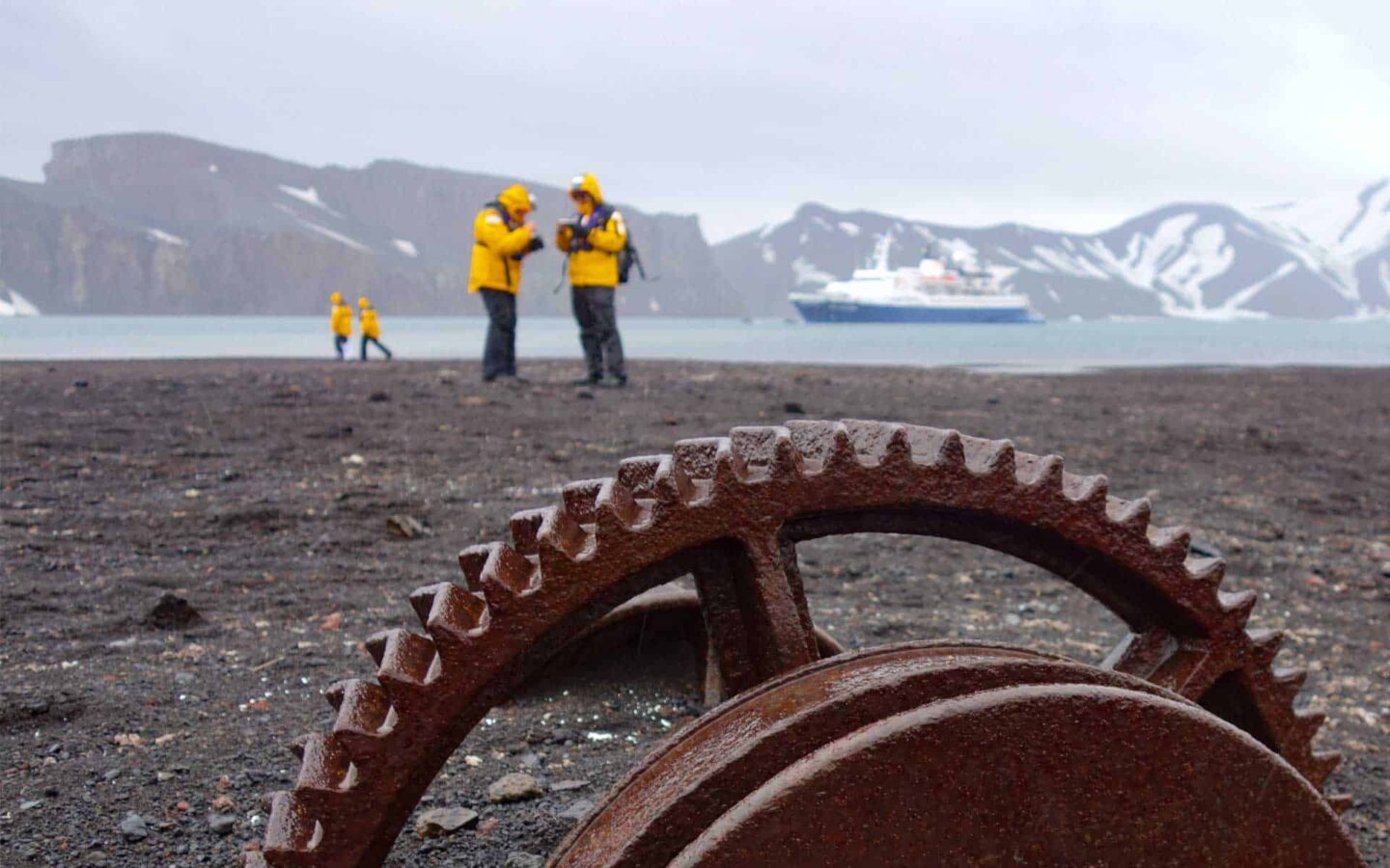 two men in yellow jackets talk in antarctica with rusty equipment in the foreground and an expedition ship and large mountains in the background