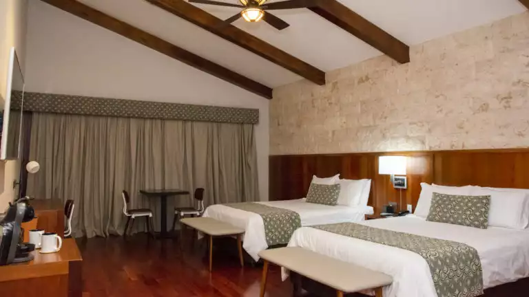 A guest room with two twin beds, wooden beams, a ceiling fan, drapes, and a television at the family-owned Los Brezos Hotel, near the Baru Volcano in Panama