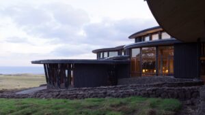 The windows and modern architecture of Posada de Mike Rapu, a LEED-certified hotel, jut above a swath of green land under a cloudy sky.