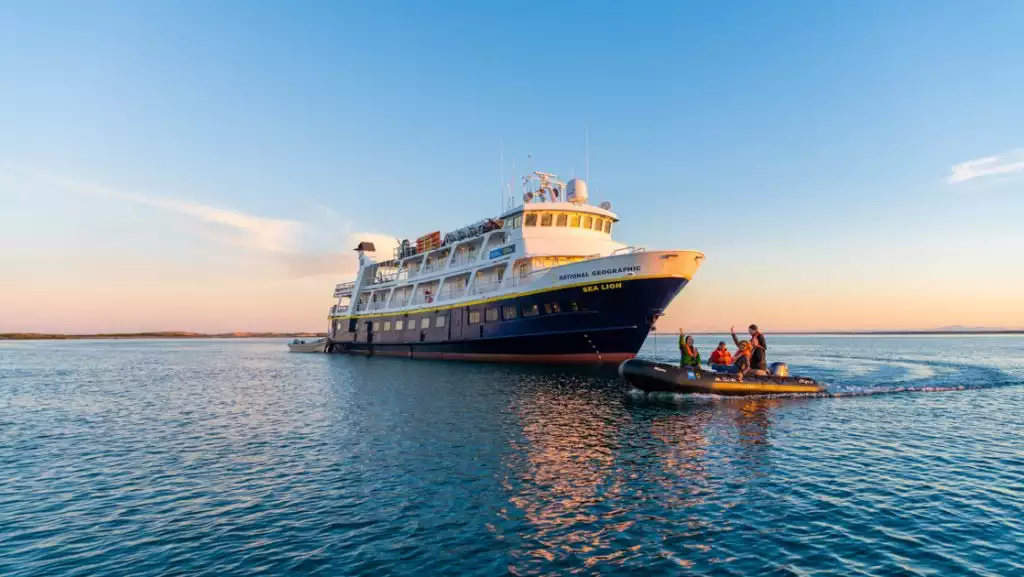 National Geographic Sea Lion small ship with dark blue hull & white upper decks sits on anchor as Zodiac cruises by at dusk.