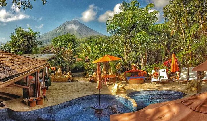 The pool, with umbrellas and lounge chairs, is set in a stone patio on the edge of the jungle with a view of the volcano at Nayara Hotel, Spa & Gardens, a luxury boutique hotel in Costa Rica