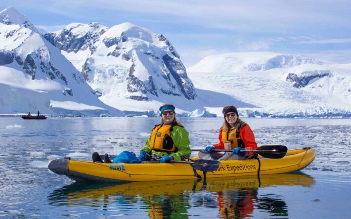 two women in a yellow kayak and brightly colored outfits sit on calm antarctica water with snowy mountains in the distance on a sunny day