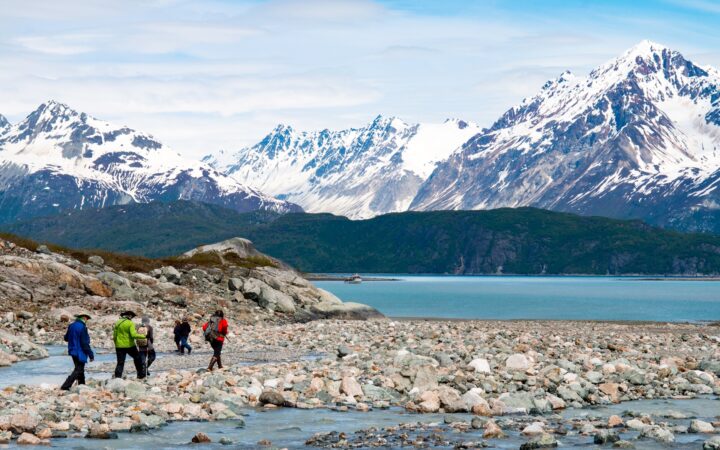 As part of an Alaska cruise shore excursion a group walks along the rocky shoreline surrounded by snow capped mountain range