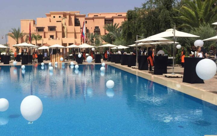 an african hotel with a large pool decorated with white balloons in the foreground and tents on the right on a sunny day