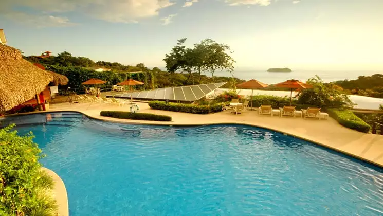 Solar panels are installed next to a pool, with a patio, umbrellas, lounge chairs, and an ocean view at Si Como No Resort on Costa Rica's Pacific Coast
