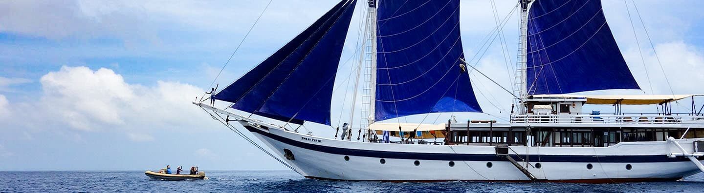 A wooden Indonesia small cruise ship is shown on starboard side with blue sails up with blue ocean, blue sky and white clouds