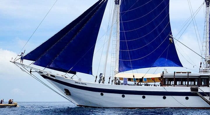 A wooden Indonesia small cruise ship is shown on starboard side with blue sails up with blue ocean, blue sky and white clouds