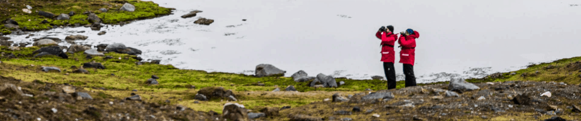 arctic travel partners stand next to each other looking through binoculars with a pond behind them