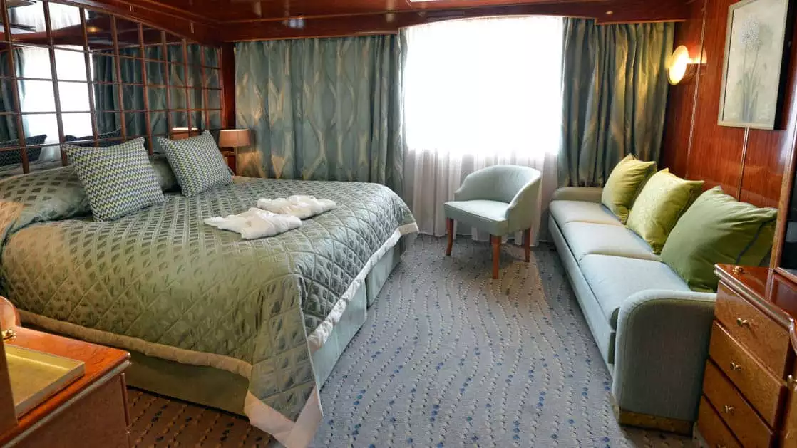 Hebridean Sky Promenade suite with large picture window, queen bed, couch, chair and desk.