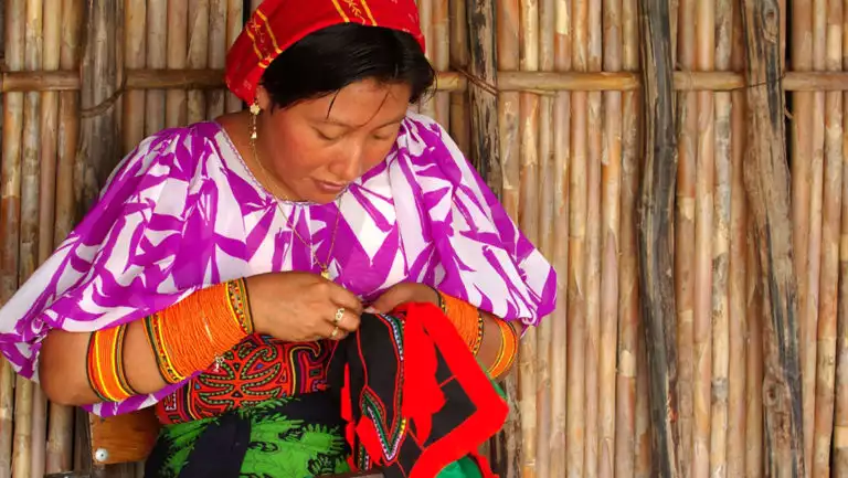 an indigenous woman in a bright top weaving in front of a thatched wooden wall on the best of panama land tour