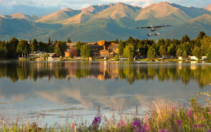 At dusk, a plane flies above the Lakefront Anchorage Lodge, with mountains in the background and water reflecting trees in the foreground.