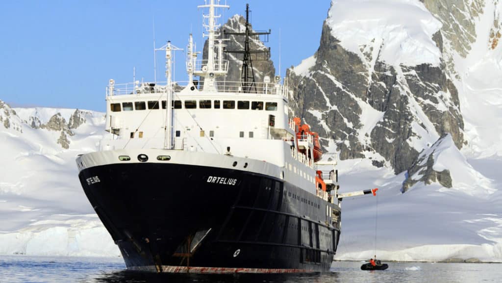 the ortelius small ship sailing with ice around it and land in the background on a sunny day
